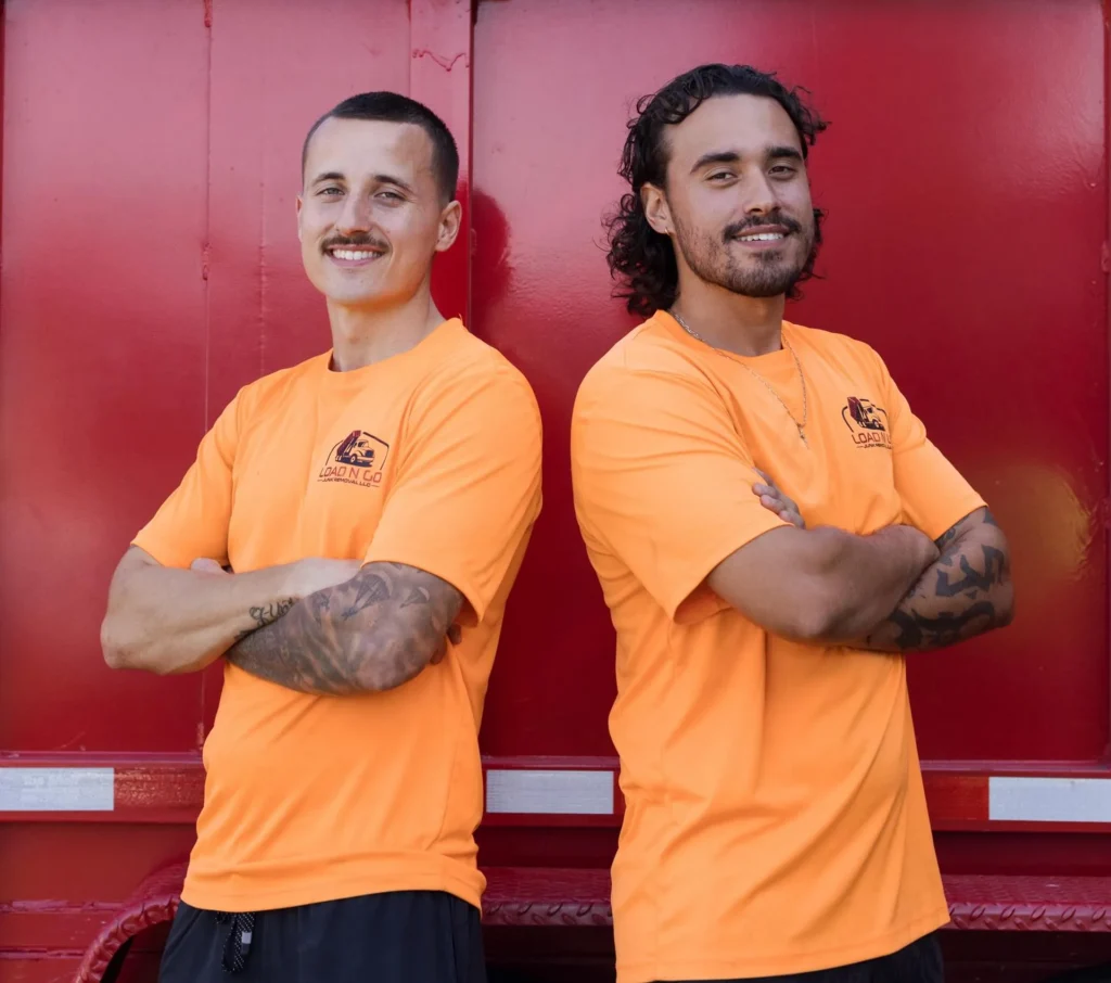 Two men in orange "Load N Go" t-shirts standing confidently with arms crossed, against a red background, representing the construction debris removal service.